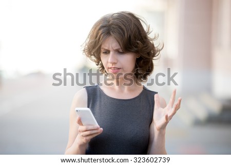 Portrait of young woman holding cellphone in hands on the street in summer, looking at screen with irritated expression, made a mistake or annoyed by texts and calls - stock photo