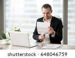 Small photo of Smiling businessman looking at document in hands while sitting at desk with laptop. Happy entrepreneur reading letter with good news. Office worker guy gets pay raise, premium bonus from his employer
