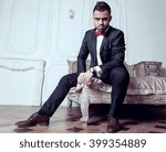 Small photo of Handsome guy with beard, side whiskers and mustache sits on a bed dressed in black suit, white shirt, red bow-tie and brown leather shoes