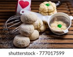 Small photo of Six lemon crinkle cookies stacked in the form of a pyramid and some Aunt Birdie's cookies with a green cherry. It is displayed with a sieve, whisk, red heart and castor sugar on a wooden board