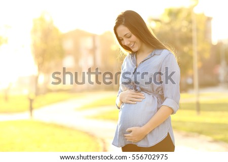 Portrait of a happy and proud pregnant woman looking at her belly in a park at sunrise with a warm back light in the background
