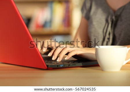 Close up of girl hands writing in a red laptop on a table at home with a warm light and a colorful background