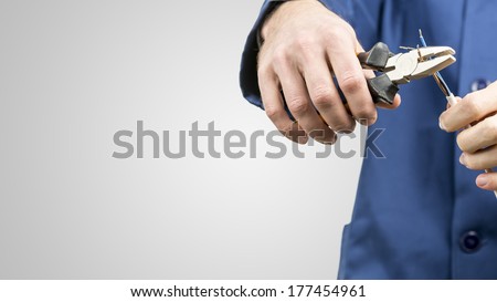 Workman or electrician repairing an electrical cable with a pair of pliers to restore supply to the house, close up view of his hands in blue overalls on grey with copyspace
