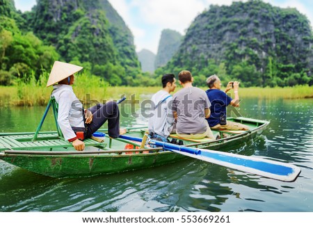 Tourists traveling in small boat along the Ngo Dong River and taking picture of the Tam Coc, Ninh Binh, Vietnam. Rower using her feet to propel oars. Landscape formed by karst towers and rice fields.