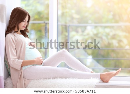 Young beautiful pregnant woman sitting on windowsill at home