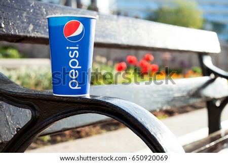 Minsk, Belarus, may 19, 2017: paper cup of Pepsi on a bench in the city on a blurry background of red flowers in the color of the logo Pepsi. Pepsi is a carbonated soft drink produced PepsiCo.