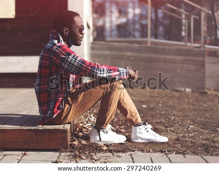 Lifestyle fashion portrait of stylish young african man listens to music evening and enjoys sunset, wearing a hipster plaid red shirt and sunglasses sitting in profile outdoors