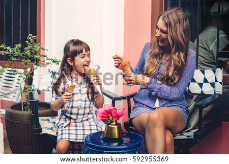 Mom with her 6 years old daughter walking along city street and eating ice cream in front of the outdoor cafe. Good relations of parent and child. Happy moments together.