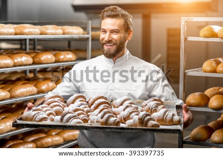 Handsome baker in uniform holding tray full of freshly baked croissants at the manufacturing