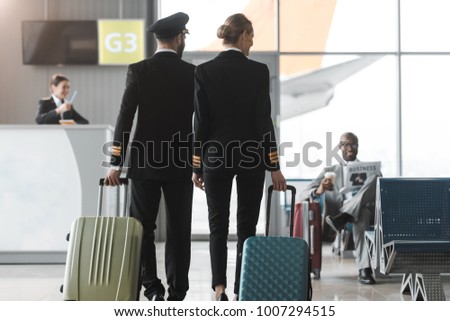 back view of male and female pilots walking by airport lobby with suitcases