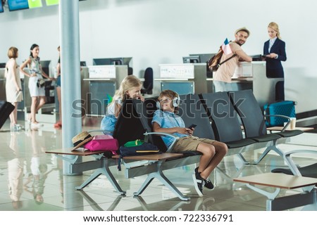 boy in headphones using smartphone with sister near by and father at check in desk at airport