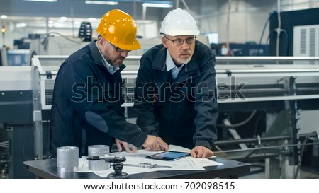 Two engineers discuss a blueprint while checking information on a tablet computer in a factory.