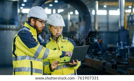 Male and Female Industrial Engineers in Hard Hats Discuss New Project while Using Laptop. They Make Showing Gestures.They Work in a Heavy Industry Manufacturing Factory.