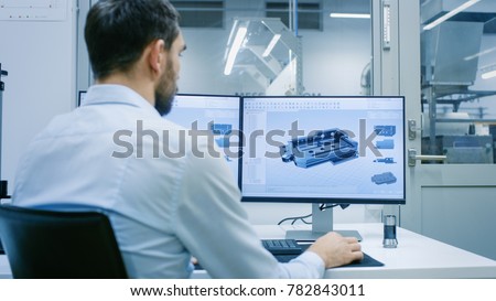Engineer/ Technician Working on a Personal Computer with Two Displays, He's Designing New Component in CAD Program. Out of the Office Window Components Manufacturing Factory is Seen.