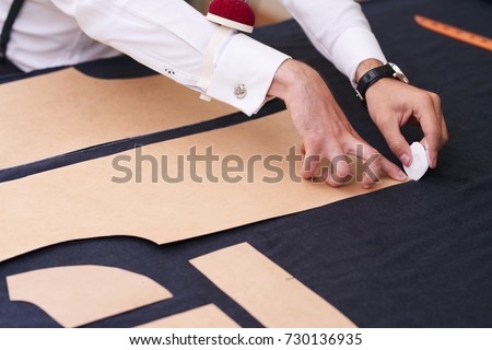 Closeup of tailors table with male hands tracing fabric making pattern for clothes in traditional atelier studio