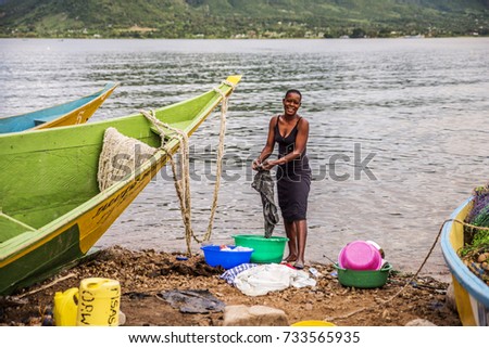 2017 May 14:Fishing village,Takawiri Island,Lake Victoria,Kenya.Water’s edge.Boats beached after fishing.African woman standing, hand washing clothes in bucket. Clean cooking dishes stacked in bucket.