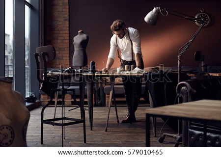 Handsome serious tailor in white shirt with brown leather suspenders working near wooden table with threads, apron and scissors in amazing atelier with antique furniture and mannequin on background