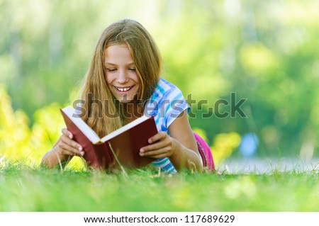 Beautiful smiling teenage girl in blue blouse lying on grass and read book, against green of summer park.