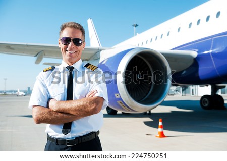 Confident pilot. Confident male pilot in uniform keeping arms crossed and smiling with airplane in the background