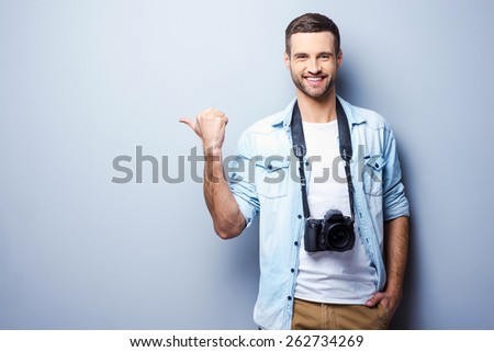 Photographers choosing it. Handsome young man with digital camera pointing away and smiling while standing against grey background
