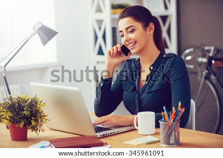 Sharing good business news. Attractive young woman talking on the mobile phone and smiling while sitting at her working place in office and looking at laptop