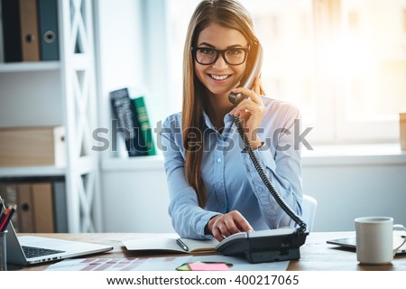 I will connect you in one second! Cheerful young beautiful woman in glasses talking on the phone and looking at camera with smile while sitting at her working place