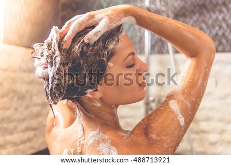 Beautiful naked young woman is smiling and using shampoo while taking shower in bathroom