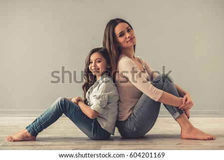 Charming little girl and her beautiful young mom are looking at camera and smiling while sitting back to back on the floor
