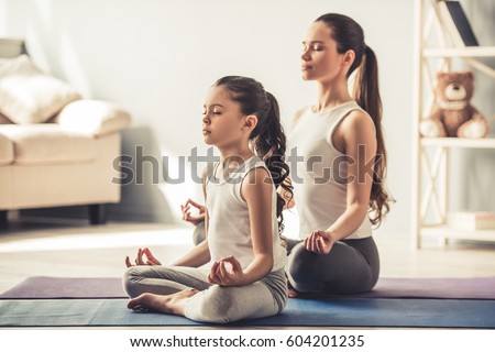 Beautiful young woman and her charming little daughter are smiling while doing yoga together at home