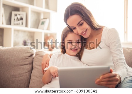 Charming little girl and her beautiful young mom are using a digital tablet and smiling while sitting on sofa at home
