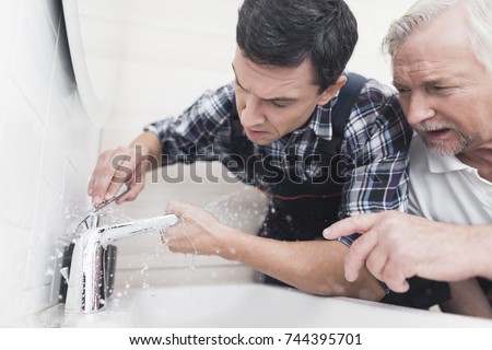 Two plumbers repair the faucet in the bathroom. One of them replaces the tap, the second one teaches the junior sanitary technician. On the young plumbers special uniform.