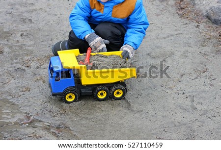 Part of the image of a child playing in the cold season on the street. A child sits near a large toy car on the wet and muddy sand.