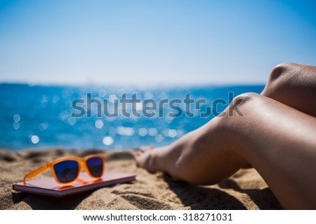 Cropped shot view of beautiful woman's legs on the beach near the sea and little red notepad on which lying fashionable colorful sunglasses, relaxing during summer weekend enjoying good day and sun