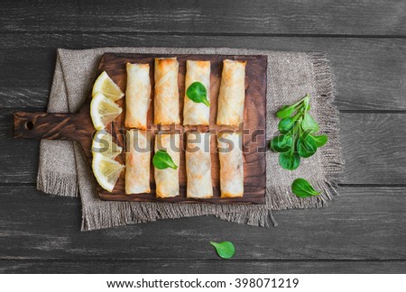 Little spring rolls on a cutting board on sacking on wooden black worn surface, lettuce, lemon slices, top view