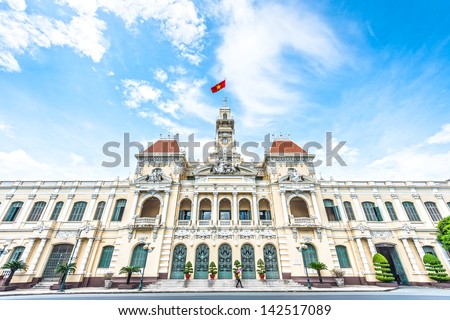 French style of building in Vietnam, Asia. Beautiful Ho Chi Minh City Hall. Facade of house with ornate design. Red flag contrasts with blue sky and clouds. Tourist attraction, famous landmark.