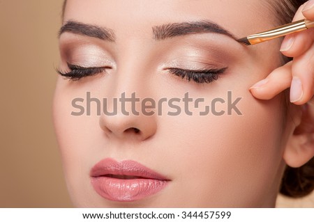 Close up of beautiful face of young woman getting make-up. The artist is applying eyeshadow on her eyebrow with brush. The lady closed eyes with relaxation