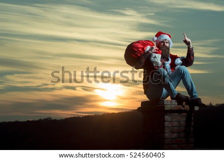 young handsome bearded bad santa claus man with long beard in checkered shirt jeans and red new year hat in christmas or xmas coat with toy sack on brick chimney in sunset, copy space