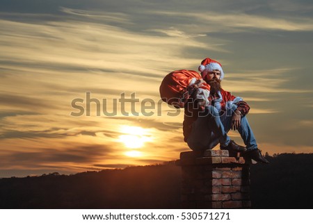young handsome bearded bad santa claus man with long beard in checkered shirt jeans and red new year hat in christmas or xmas coat with toy sack on brick chimney in sunset, copy space