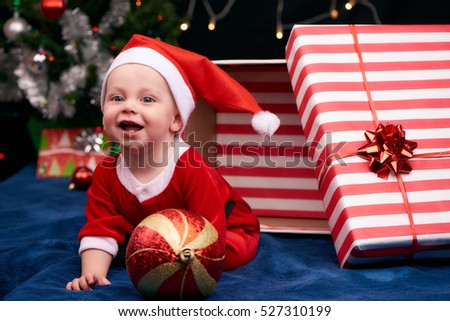 Baby boy in a santa outfit busy crawling out of a gift wrapped box as a present under the christmas tree, along with the other gifts and decorations.