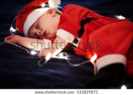 Baby boy wearing Santa outfit sleeping on blue blanket with fairy lights around him.