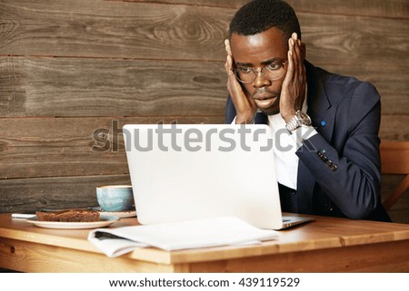 Handsome African corporate worker in glasses using laptop for distant work, sitting at the hotel lobby, looking at the camera in shock, surprised with sensational news, holding hands on his cheeks