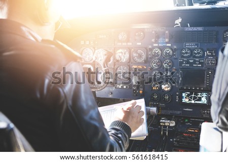 People and occupation concept. Adult captain of jet airliner wearing black leather jacket and headset sitting in front of onboard instruments, filling in safety report before departure. Blurred shot