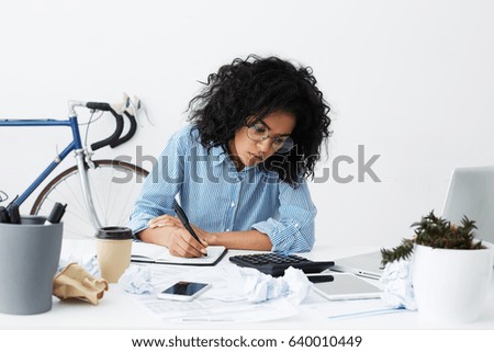 Attractive young dark-skinned female columnist suffering from writer's block, working on new article for online women's magazine, making notes in sketchbook, surrounded by crumpled paper balls