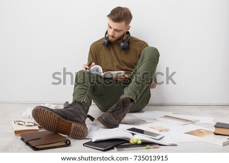 Busy male student wears casual clothes and boots, writes notes, being invlolved in studying before session. Creative young writer has inspiration, sits on floor, put down notes in diary, isolated