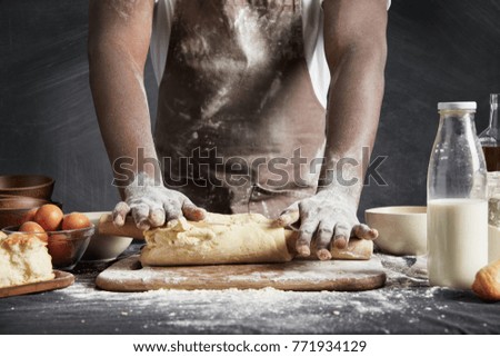Image of hard working male cook or baker with dark skin wears apron, sheets well made dough, going to bake tart, isolated over black chalk background. Unrecognizable African American chef on kitchen