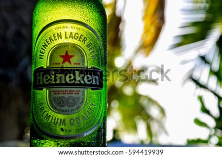 Aitutaki, Cook Islands - January 23, 2012: Close up detail of a Heineken beer bottle, illuminated by the evening sun. In the background there are palm trees on the beach of Aitutaki, Cook Islands.
