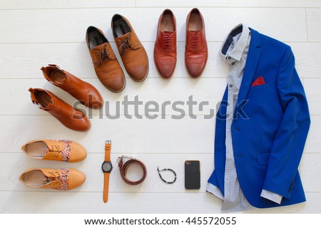 Flat lay set of brown shoes circled around men`s blue suit with shirt and some objects like watches, belt, bracelet and phone on white wooden background. Top view.