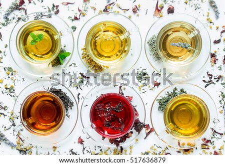 Six cups with different types of tea black, green, hibiscus, chamomile, peppermint, lavender on white background of scattered tea leafs. Six types of tea on a white background of scattered tea leafs.