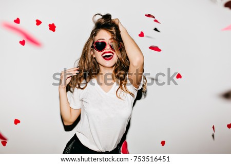 Indoor portrait of amazing caucasian female model in trendy t-shirt touching her long shiny hair. Laughing refined woman in sunglasses having fun with red confetti.