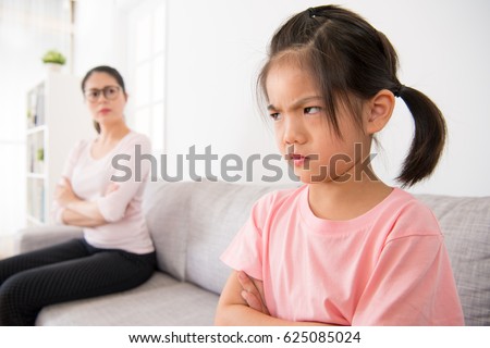 kindergarten children is scolded by school teacher and feel angry sitting in the sofa area of the classroom and the blurred teacher looked at her seriously in the background.
